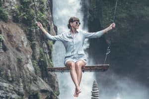 Image of joyful woman on swing with waterfall in the background as well as balanced stone piles.